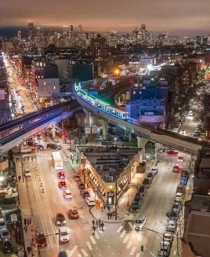 The image depicts a bustling cityscape at night, dominated by tall buildings and illuminated streets. A large, prominently lit structure resembling an elevated train or subway line arches over the street, emitting blue and green lights. Below, numerous cars and pedestrians populate the streets, which are lined with parked vehicles. Several buildings display bright signage and advertis

ements, indicating a lively urban area. In the background, a dense array of skyscrapers reveals an expansive city skyline under a dusky sky, suggestive of a major metropolitan area like New York City.