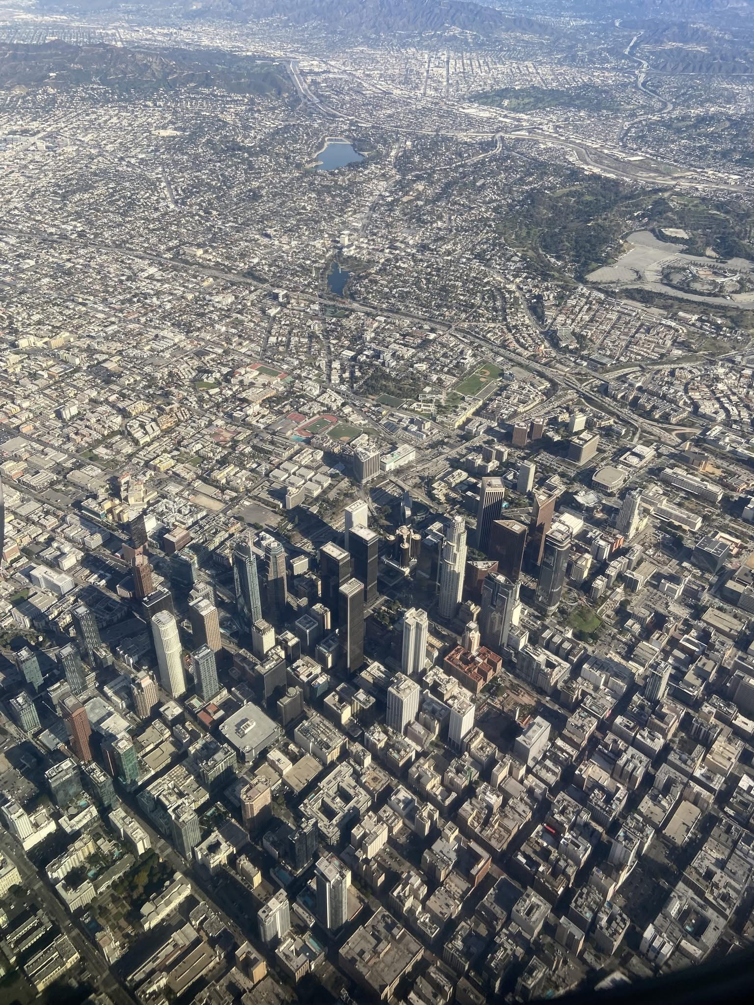 The image depicts an aerial view of a densely populated urban area with a mix of high-rise buildings and residential structures. The cityscape features a prominent cluster of skyscrapers in the center, suggesting a central business district. Surrounding the downtown area are numerous smaller buildings and a grid-like pattern of streets. There are also visible green spaces and parks scattered throughout the city. The layout and architecture suggest it could be a major metropolitan area like Los Angeles.