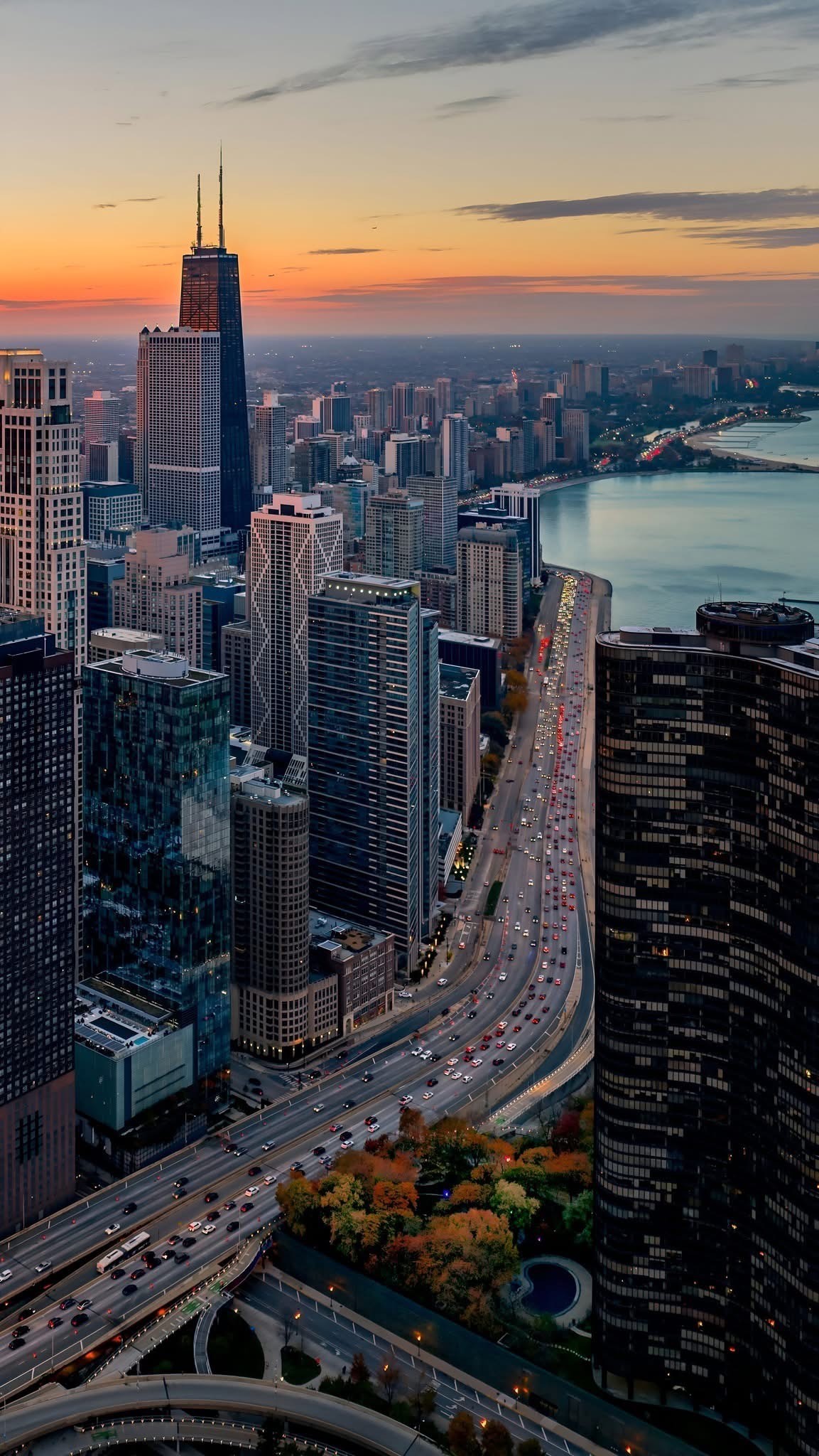 The image depicts a cityscape during sunset. The tallest building in the center is likely the John Hancock Center, suggesting the city is Chicago. The skyline is filled with numerous high-rise buildings, and a major highway runs through the foreground, bustling with traffic. The Chicago River is visible on the right, reflecting the setting sun's warm hues. The sky is painted with shades of orange and pink, creating a picturesque urban scene.